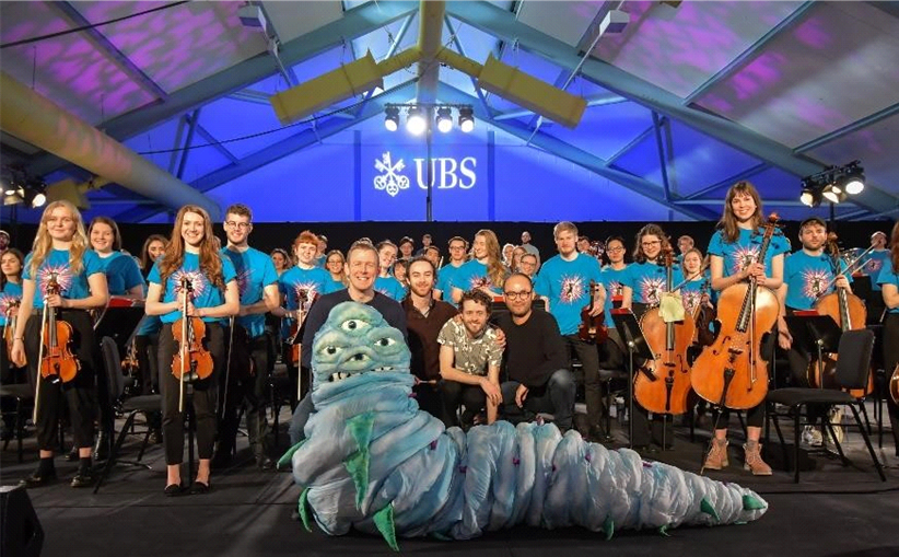 A large group of Royal Welsh College of Music & Drama performers stood on stage holding string instruments and smiling. Four other people are sat at the front of the stage behind a large manufactured, blue creature which looks like an alien worm with four eyes.