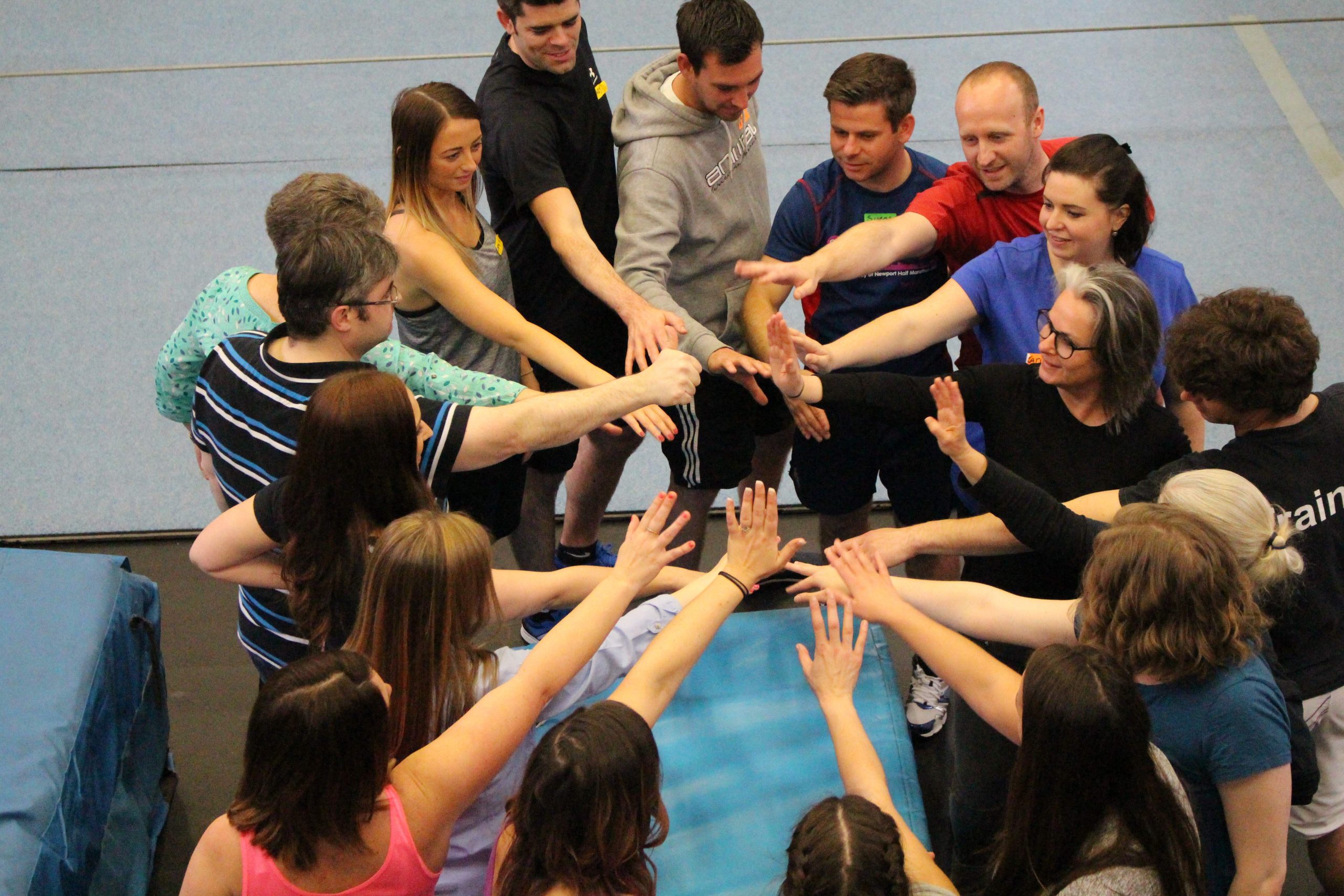 A group of people, dressed in sportswear, stand in a circle at NoFit State Circus with each individual extending one arm into the centre of the circle. 