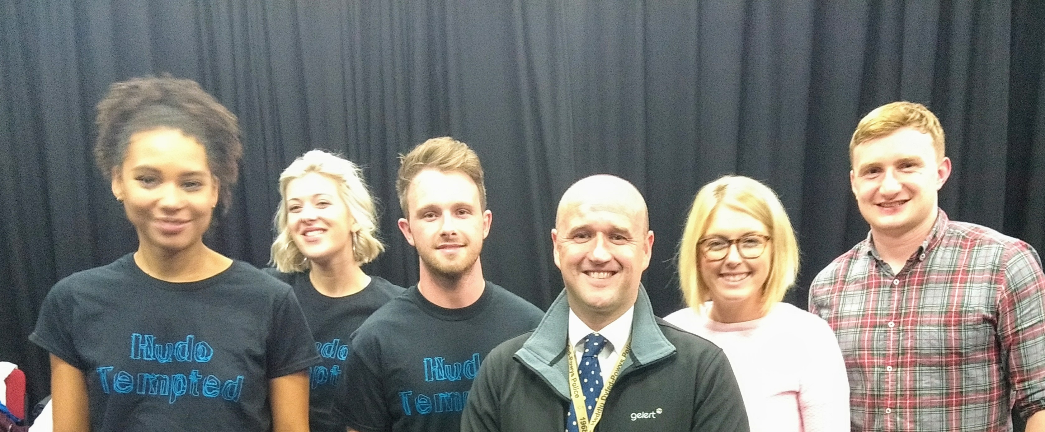 Six people stand in a row in front of long black curtains, smiling at the camera. 