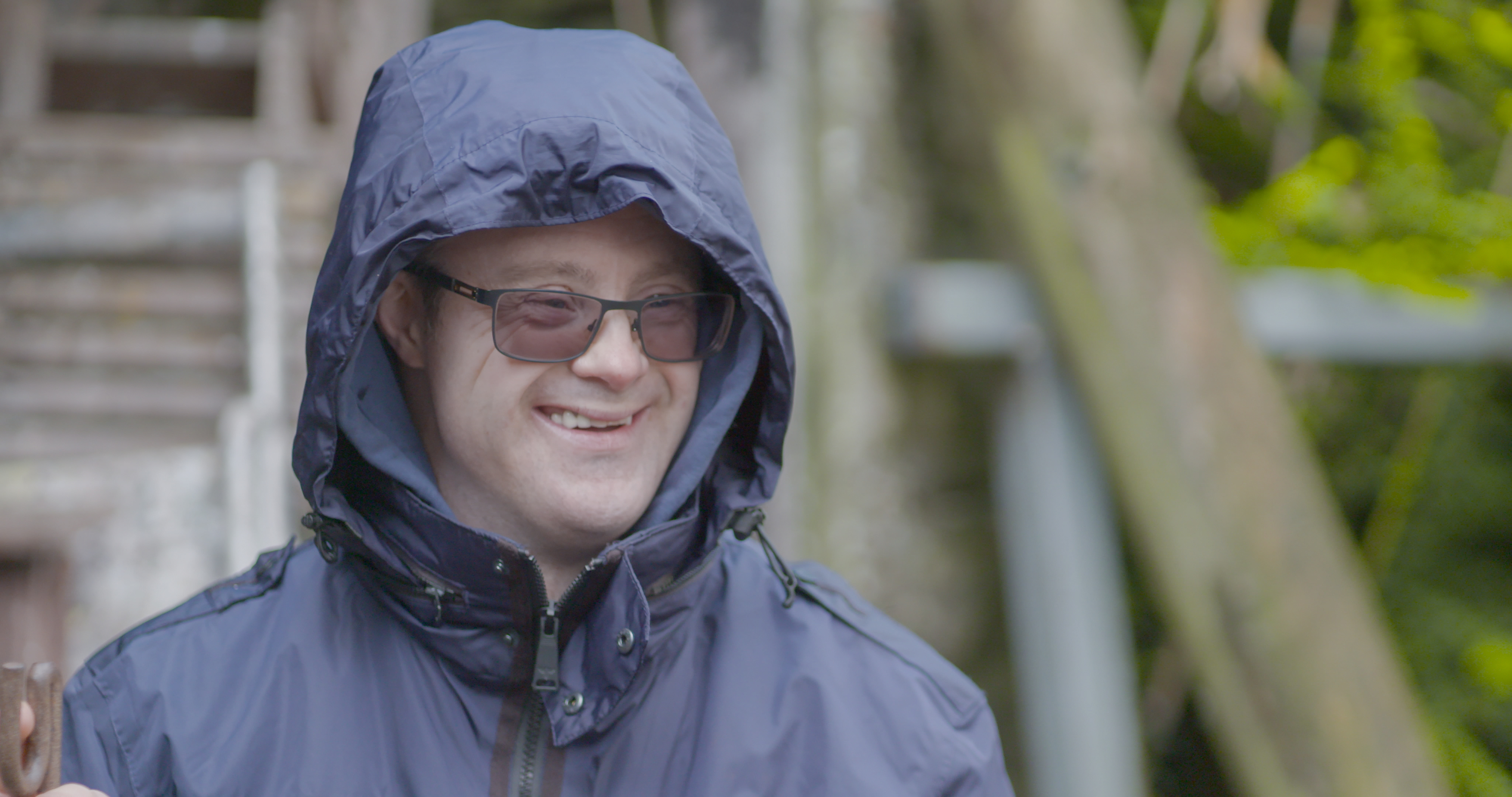 A smiling Hijinx actor wearing glasses and a blue hooded coat stands in front of a blurred background of a wooden structure and green branches.