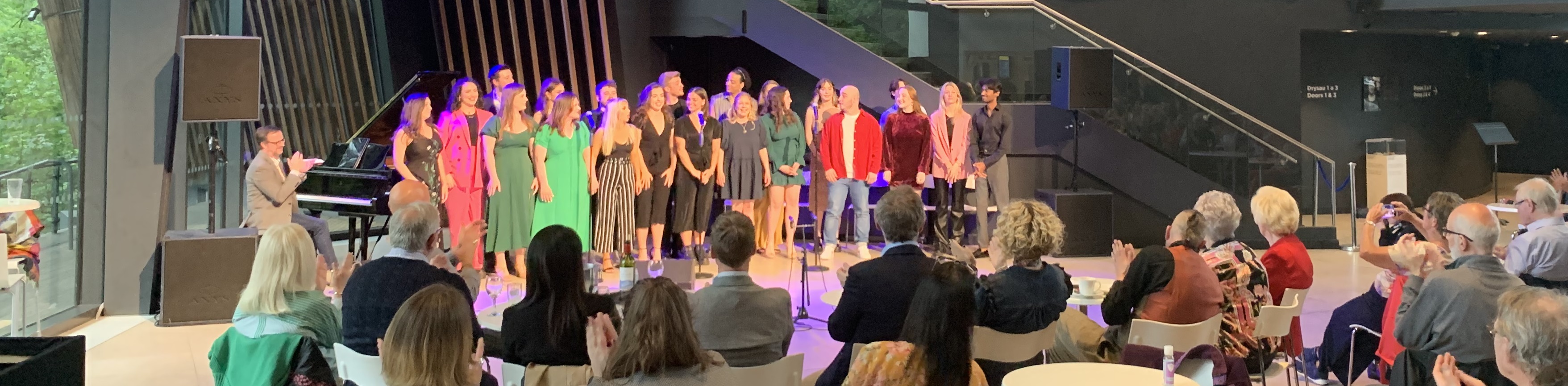 Twenty one Royal Welsh College of Music and Drama singers and one pianist on stage in front of a clapping audience. 