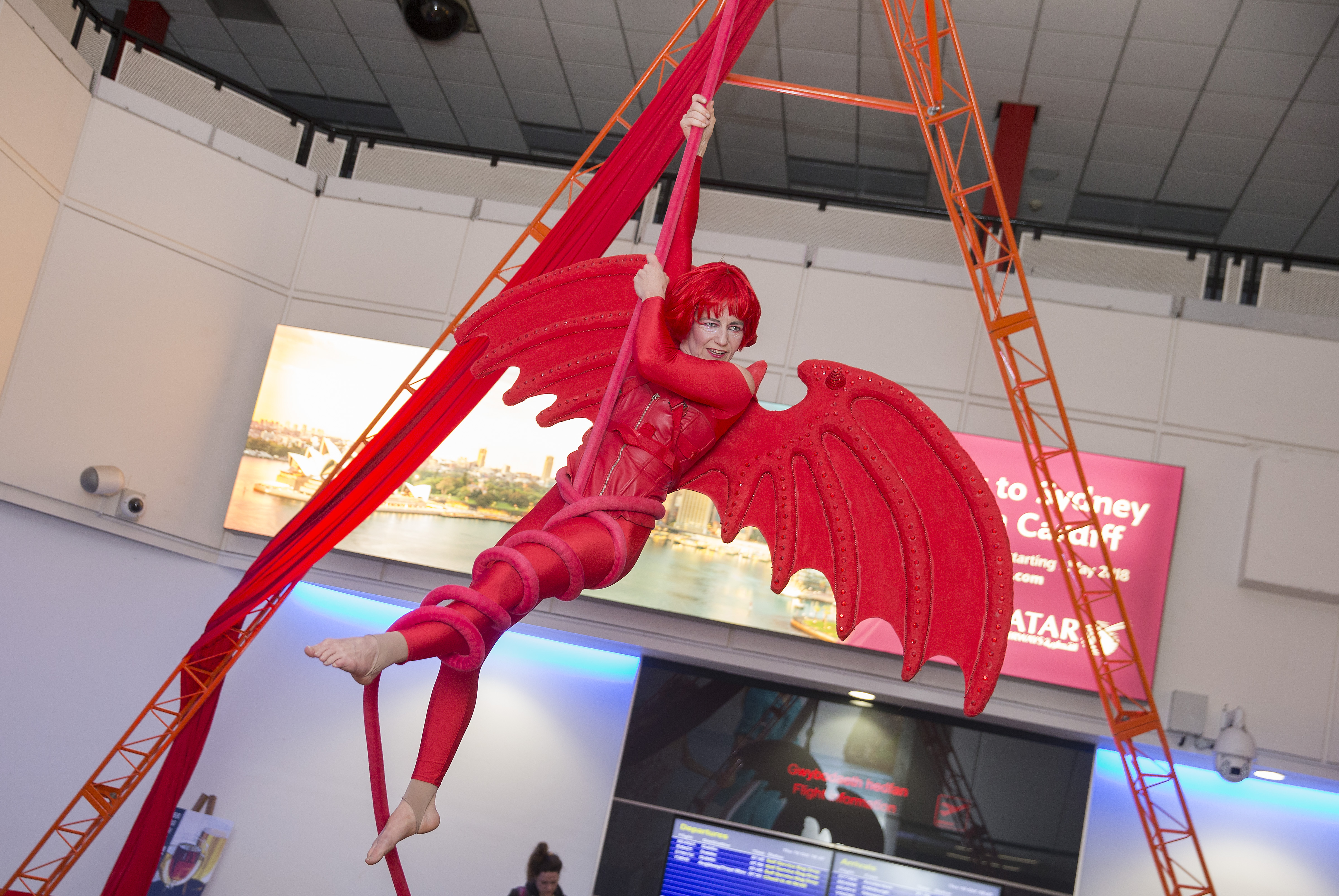 An Organised Kaos circus performer dressed as a red dragon suspended from a rig at Cardiff Airport departures gate