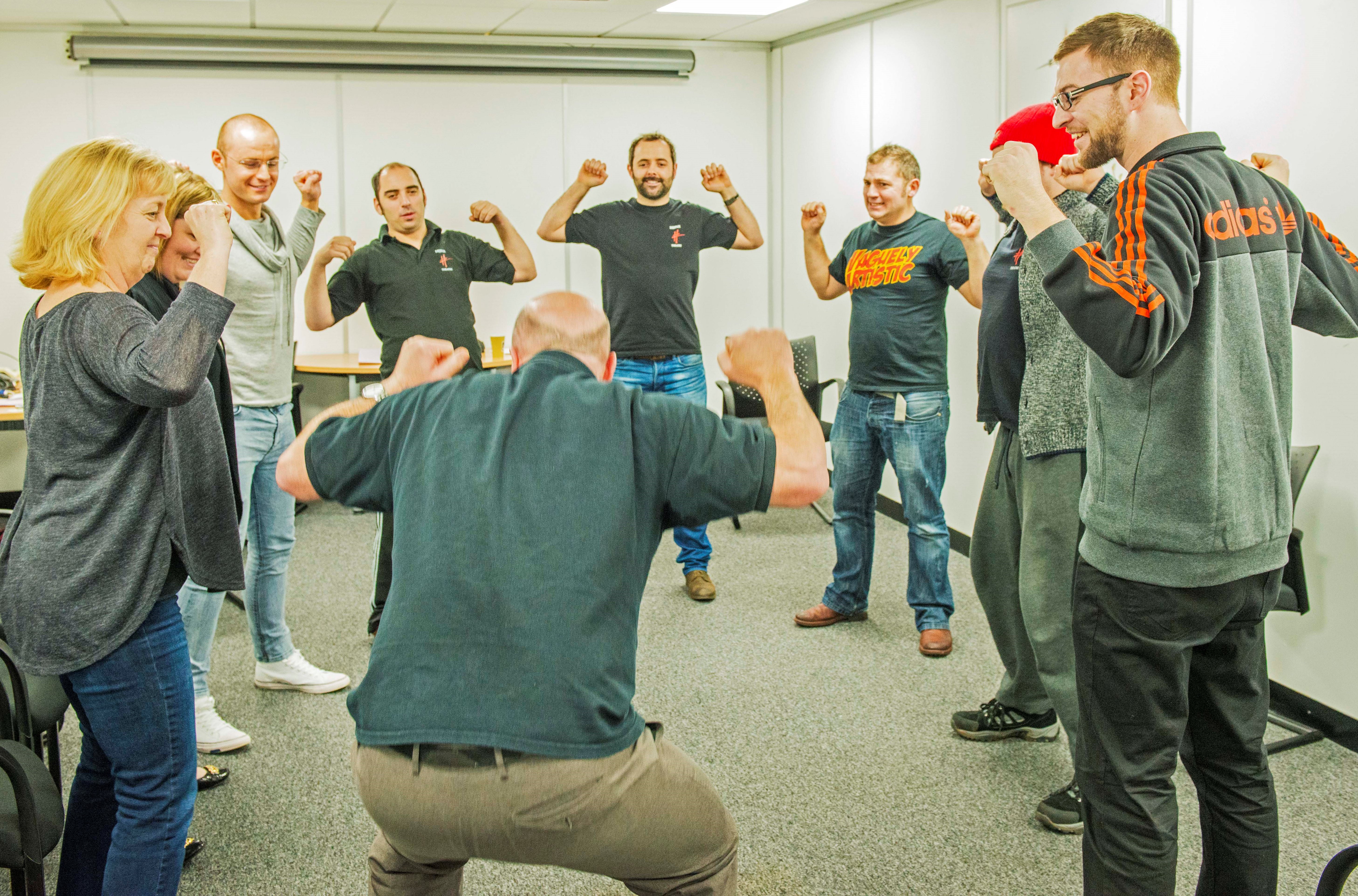 Nine people stand in a circle with arms raised and fists balled, taking part in an arts-based training activity with Hijinx Theatre