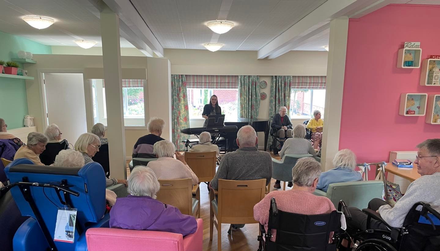 A group of care home residents watching a person singing and playing the keyboard.