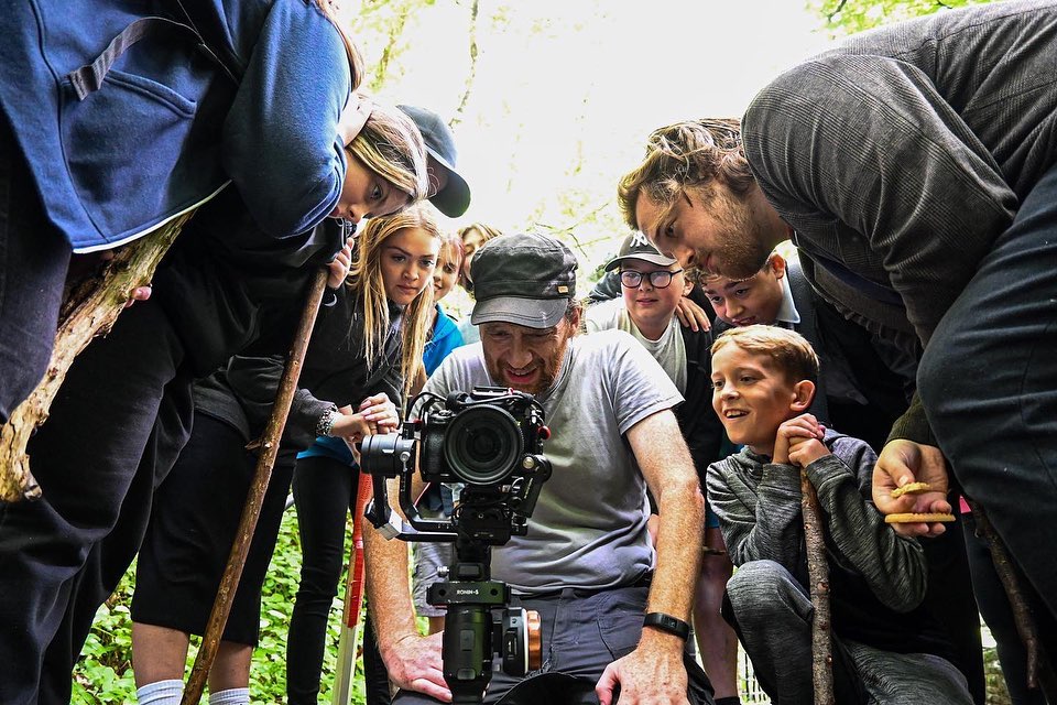 A group of people of different ages crouch down behind around a camera to look through the viewfinder, on the set of an It’s My Shout production 