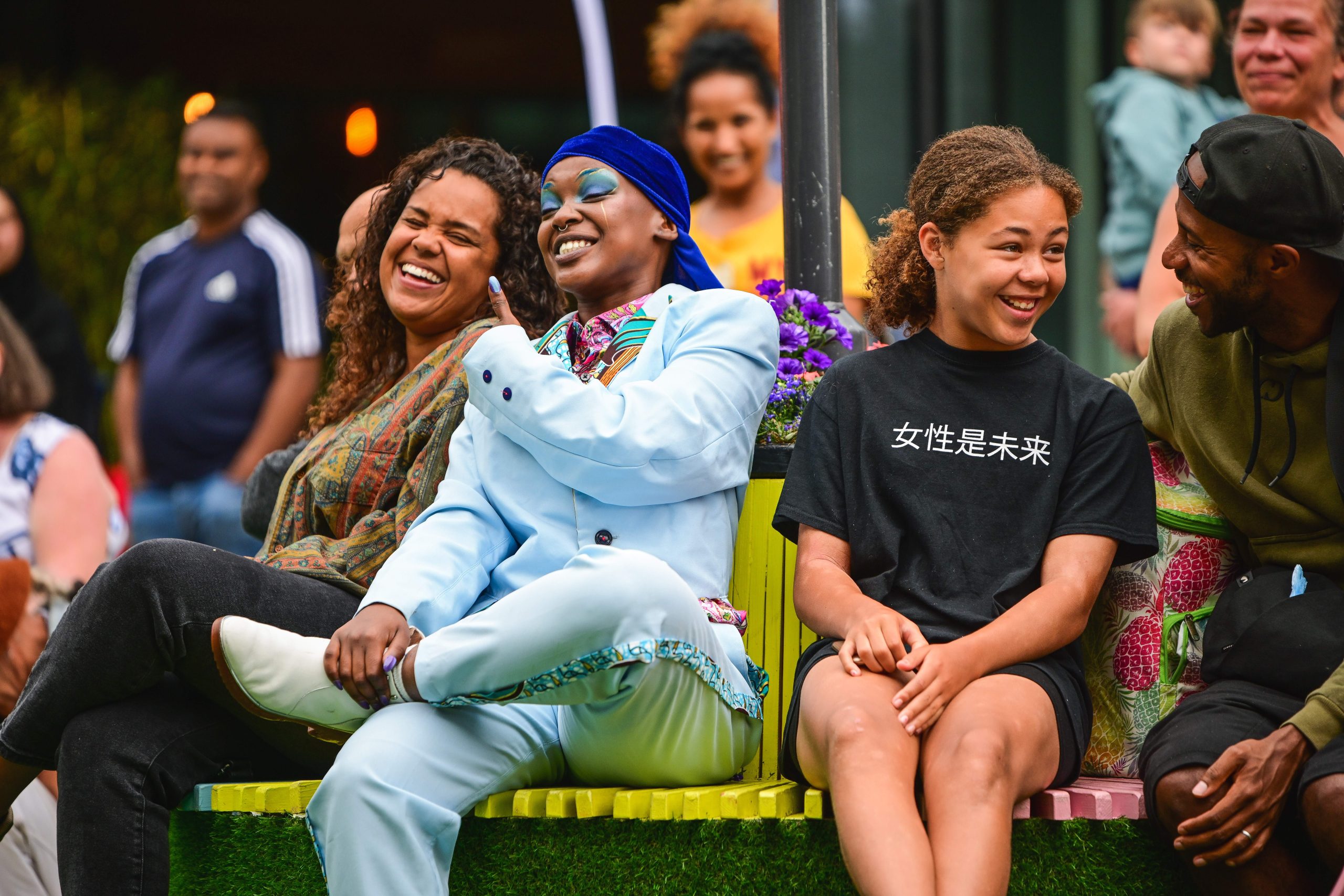 Four people, including a performer, sat on a multi-coloured bench in the street, laughing.