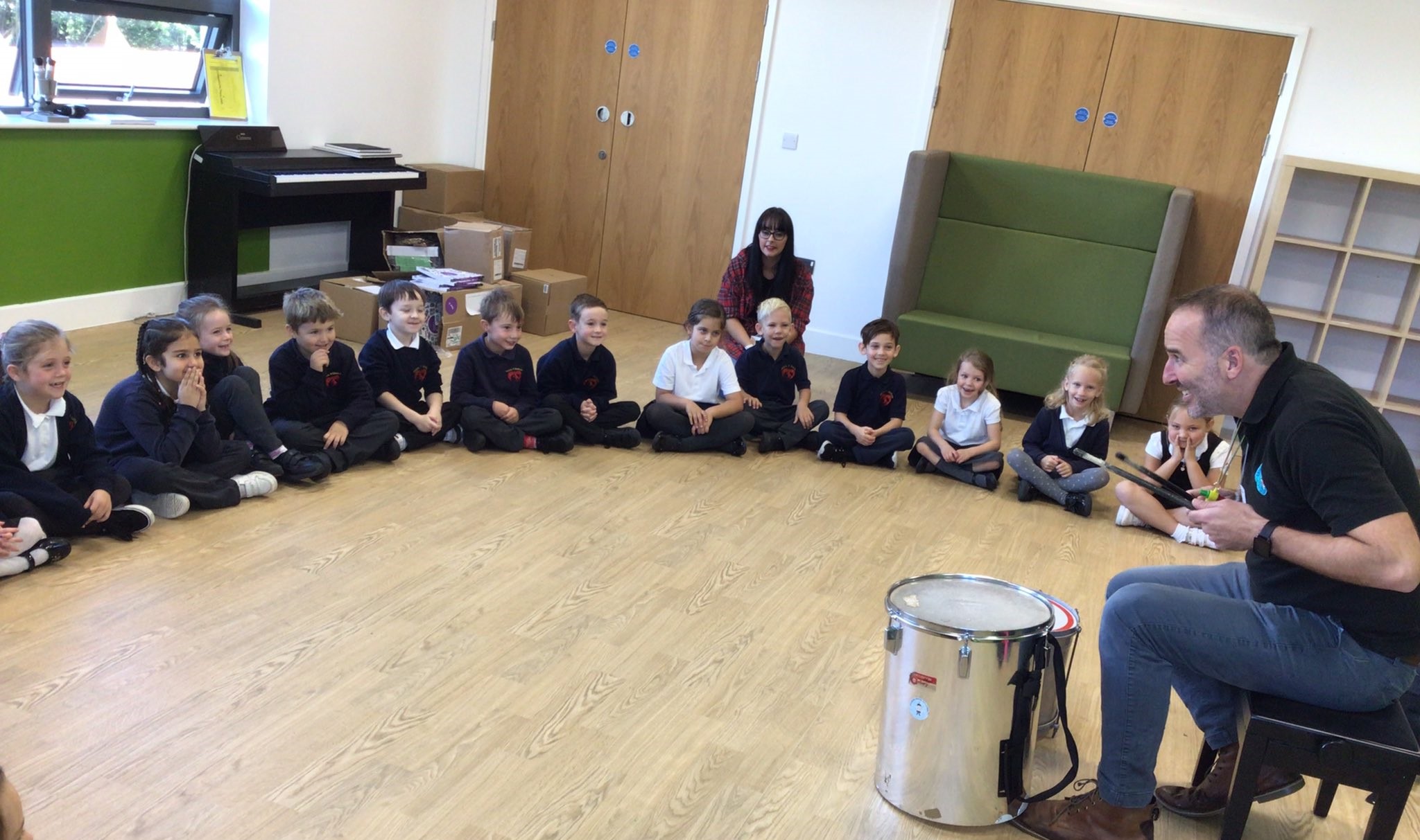 A group of young school children smiling and sat in a semi-circle on a wooden floor, watching a performer play the drums.