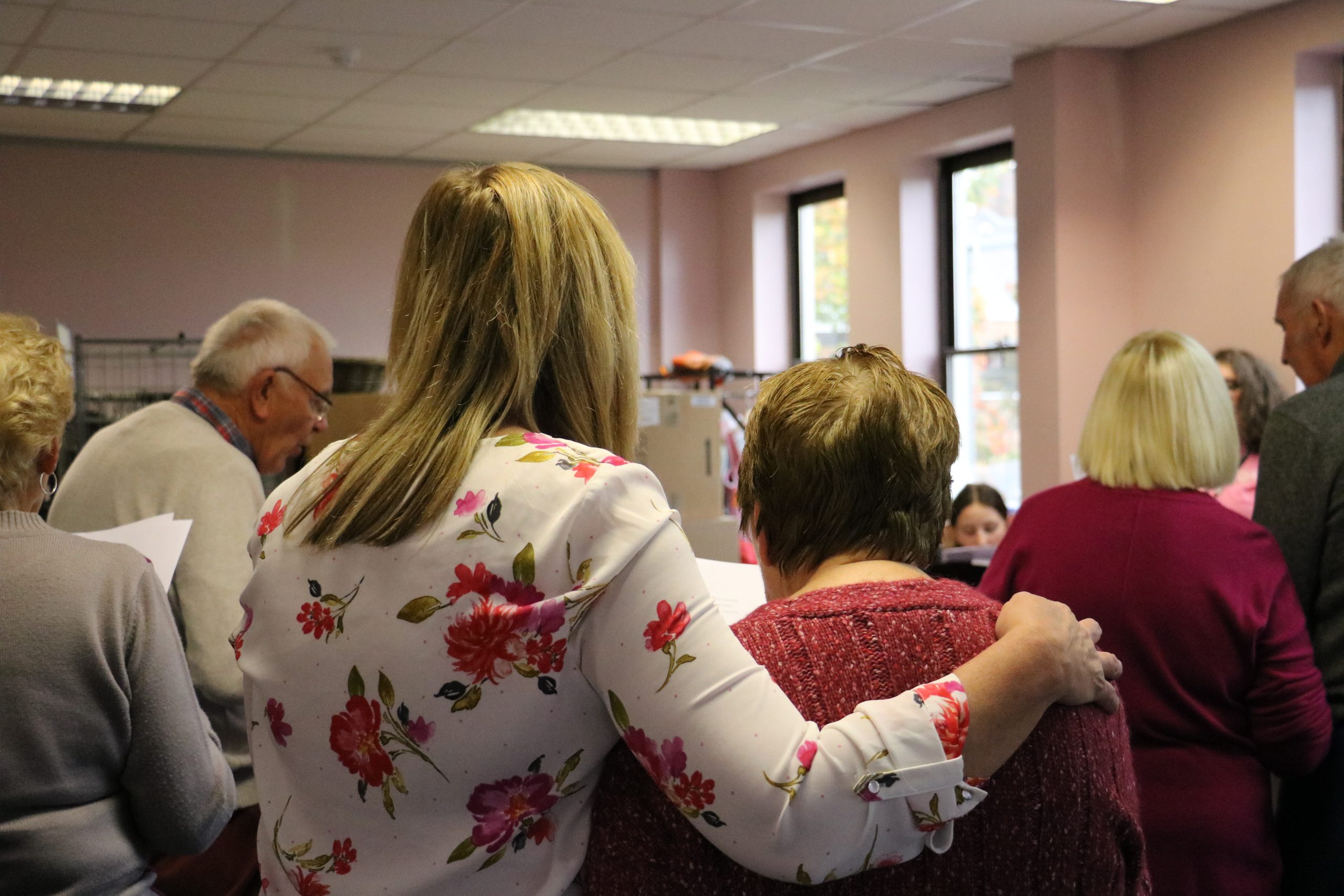 A group of Memory Arts Café participants facing away from the camera, stood in a semi-circle, singing.  