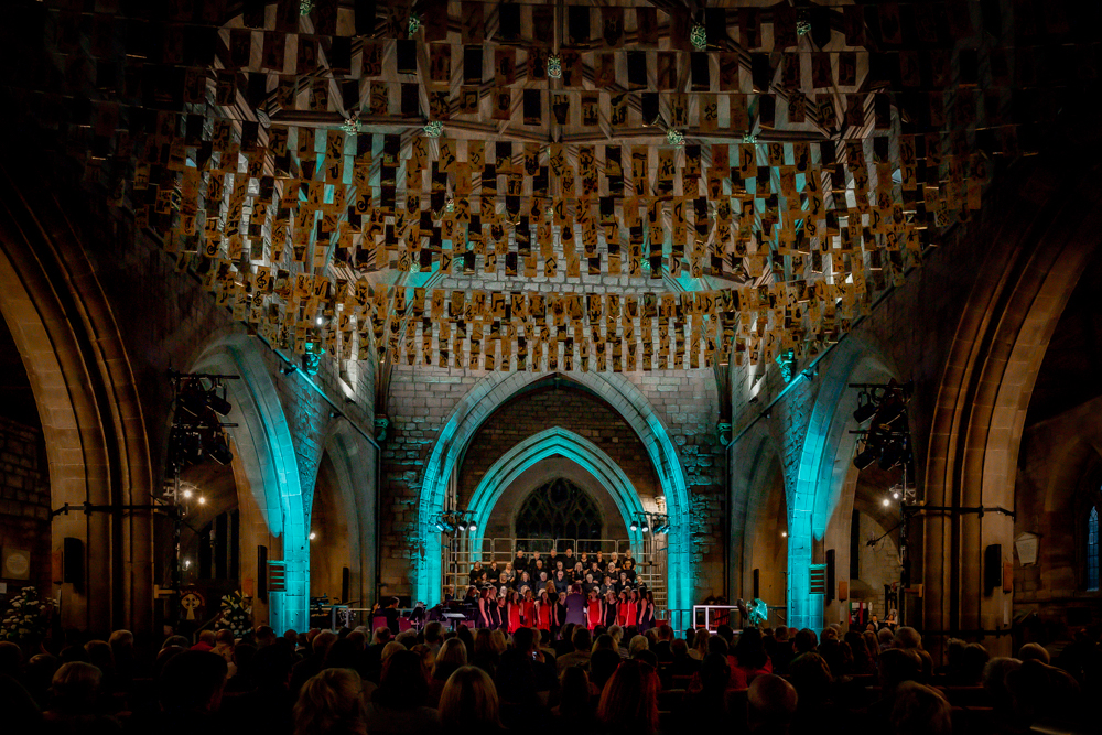 A group of performers on stage in a cathedral in front of a large audience.  Many rows of gold bunting hangs at ceiling height across the interior of the cathedral.