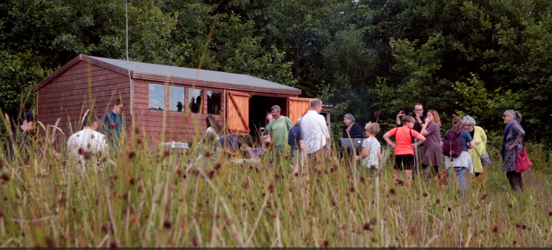 The forefront is full of strands of green grass and wild flowers. To the rear is a wooden shed with four windows and double doors, which are opened outwards. There are around fifteen people of all ages standing outside the shed with a barbecue open in the middle.  