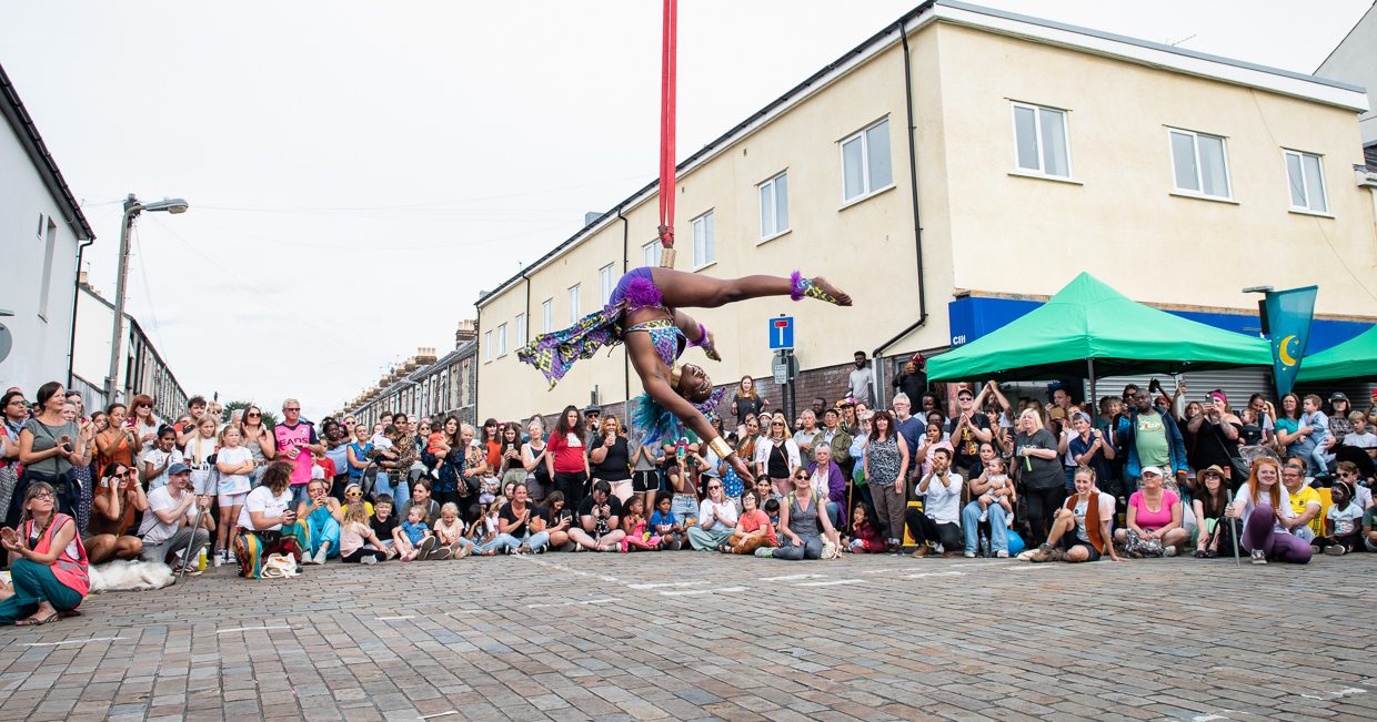 A NoFit State Circus performer hanging upside down from a circus rope in front of a large audience of people on a Cardiff street