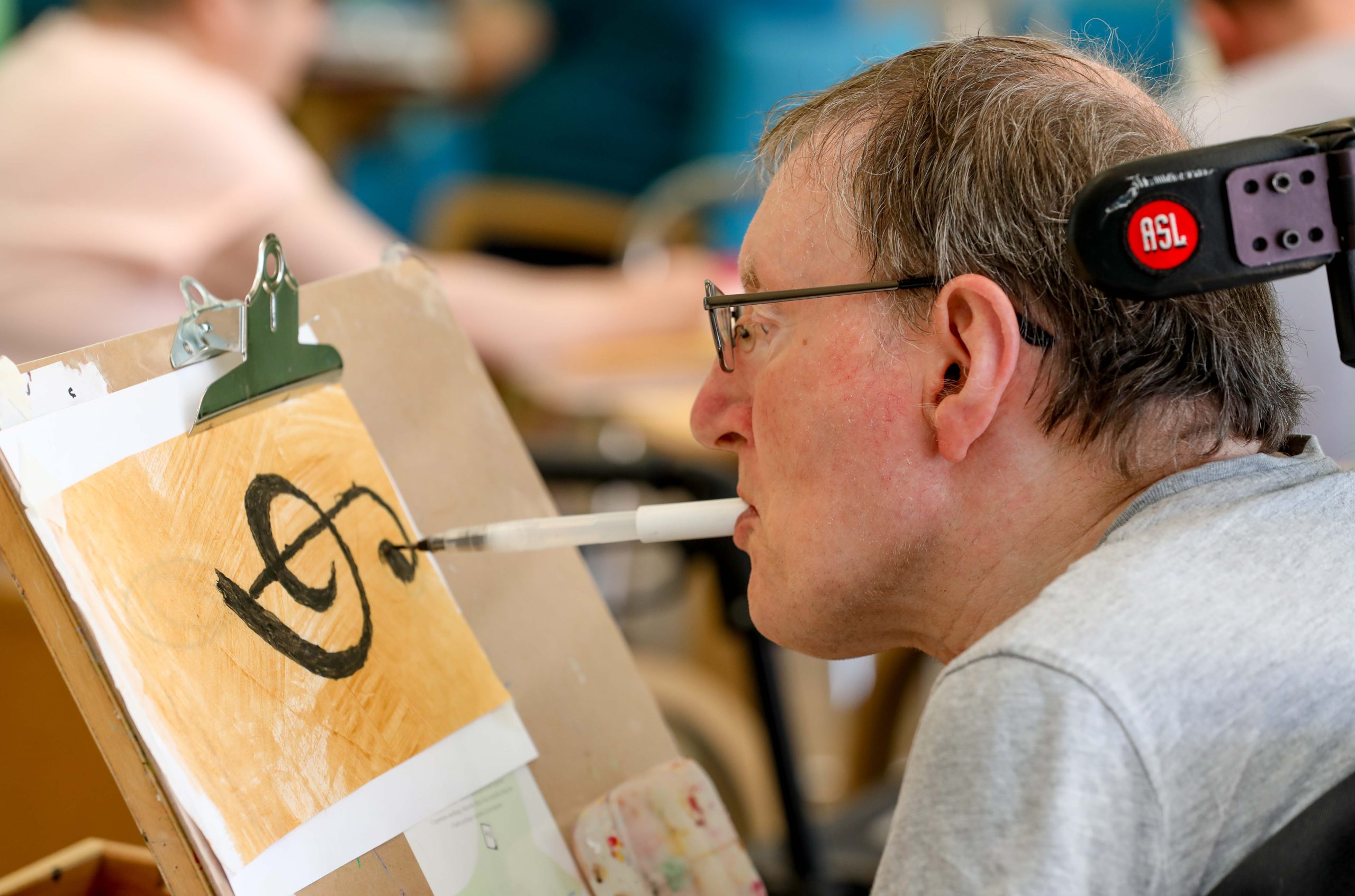 A person in a wheelchair, holding a pen in their mouth and using it to draw a music note onto a piece of paper painted gold which is clipped to an easel. 