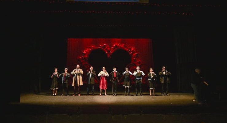 Ten people stood on a stage, wearing costumes and holding their fingers in a heart shape in front of their chests. The back of the stage is black, with a large red rectangle to the centre, with a hollow black heart in the middle. 