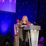 Actor Di Botcher standing behind a lectern on a presentation stage