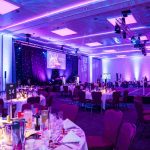 A large room full of laid tables surrounded by chairs. Stage lights on the ceiling, a presentation stage with two lecterns and a large screen, a large performance stage in the centre of the room. Dimmed lighting with pink and white downlights and up lights.